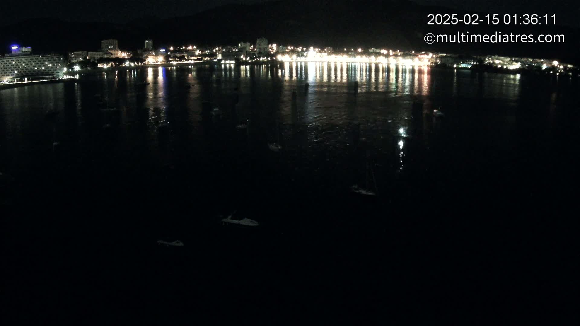 A nighttime view of a dark body of water with a few boats, reflecting the lights of a city along the shore.