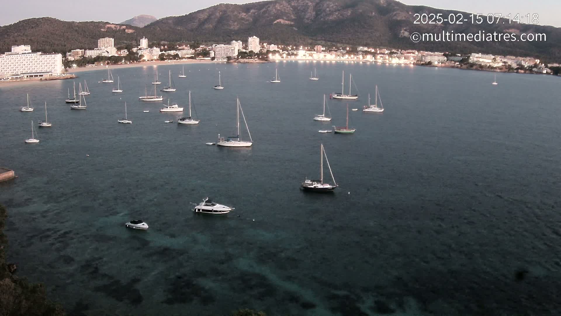 Numerous sailboats and motorboats are anchored in a calm bay near a coastline with hotels and mountains in the background under a twilight sky.