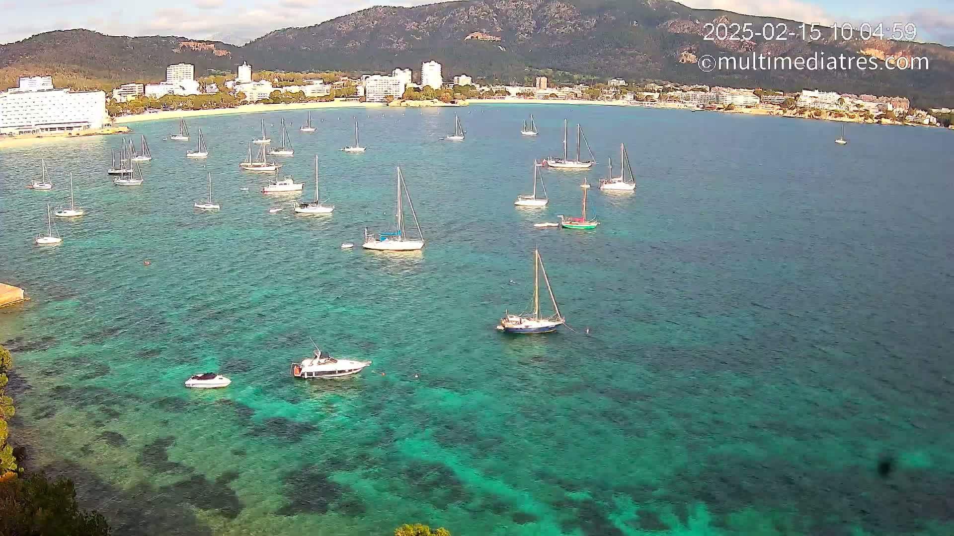 Many sailboats and motorboats are anchored in a calm, clear turquoise bay near a beach and town backed by mountains under a mostly sunny sky.