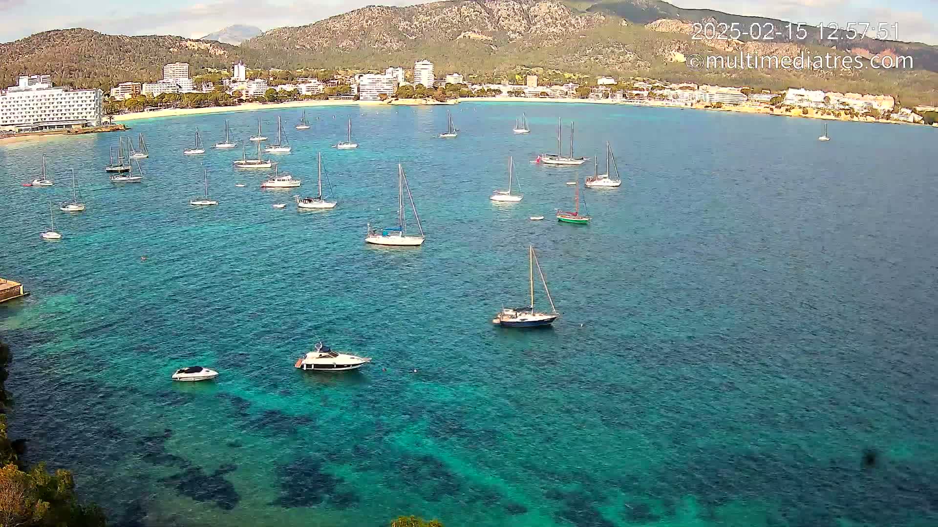 A calm, clear turquoise bay with numerous sailboats and motorboats is nestled against a shoreline of hotels and buildings backed by a mountain range under a mostly sunny sky.