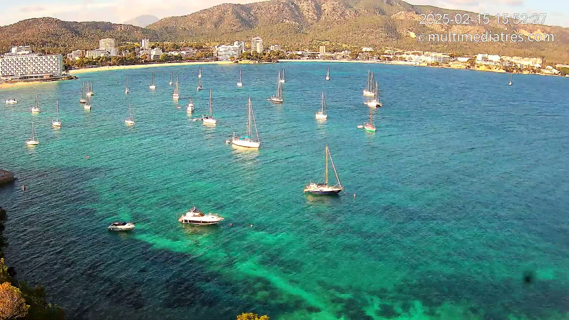 Numerous sailboats and motorboats are anchored in a calm, clear turquoise bay, with a town and mountains visible in the background under a mostly sunny sky.
