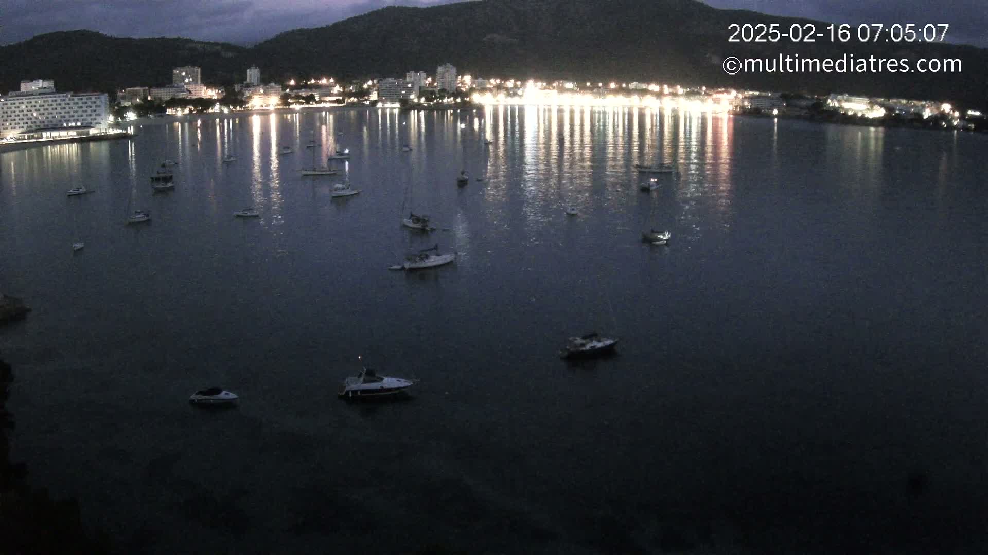 Many boats are moored in a dark, calm bay at night, with a brightly lit town visible on the far shore under a twilight sky.