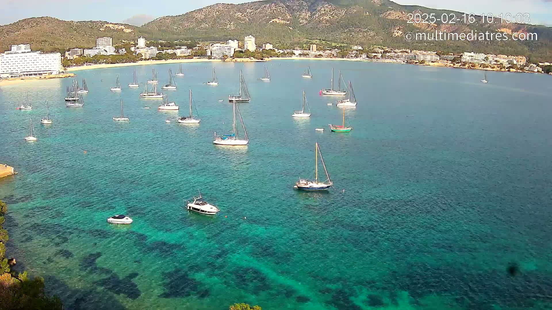 Numerous sailboats and motorboats are anchored in a calm, clear turquoise bay, with a shoreline of hotels and mountains in the background.