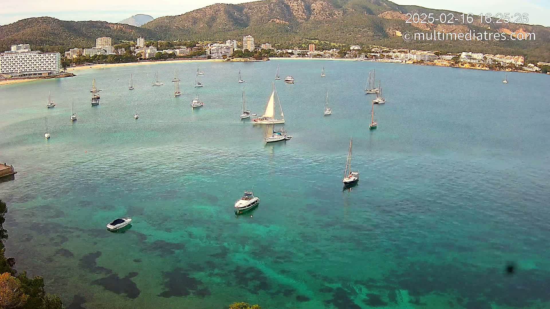 A calm, clear, teal-colored bay with numerous sailboats and motorboats anchored near a coastline featuring hotels and mountains under a mostly clear sky.