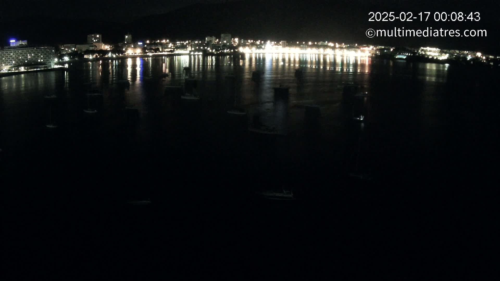 Numerous boats are moored in a dark body of water at night, reflecting the lights of a city on the opposite shore.