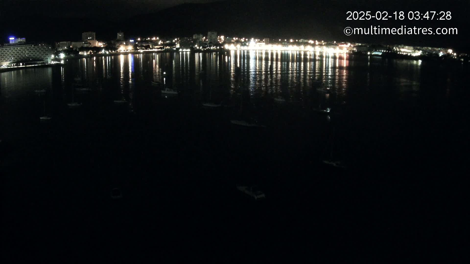 A nighttime view of a calm body of water reflecting the lights of a city skyline, with several boats scattered across the water's surface.