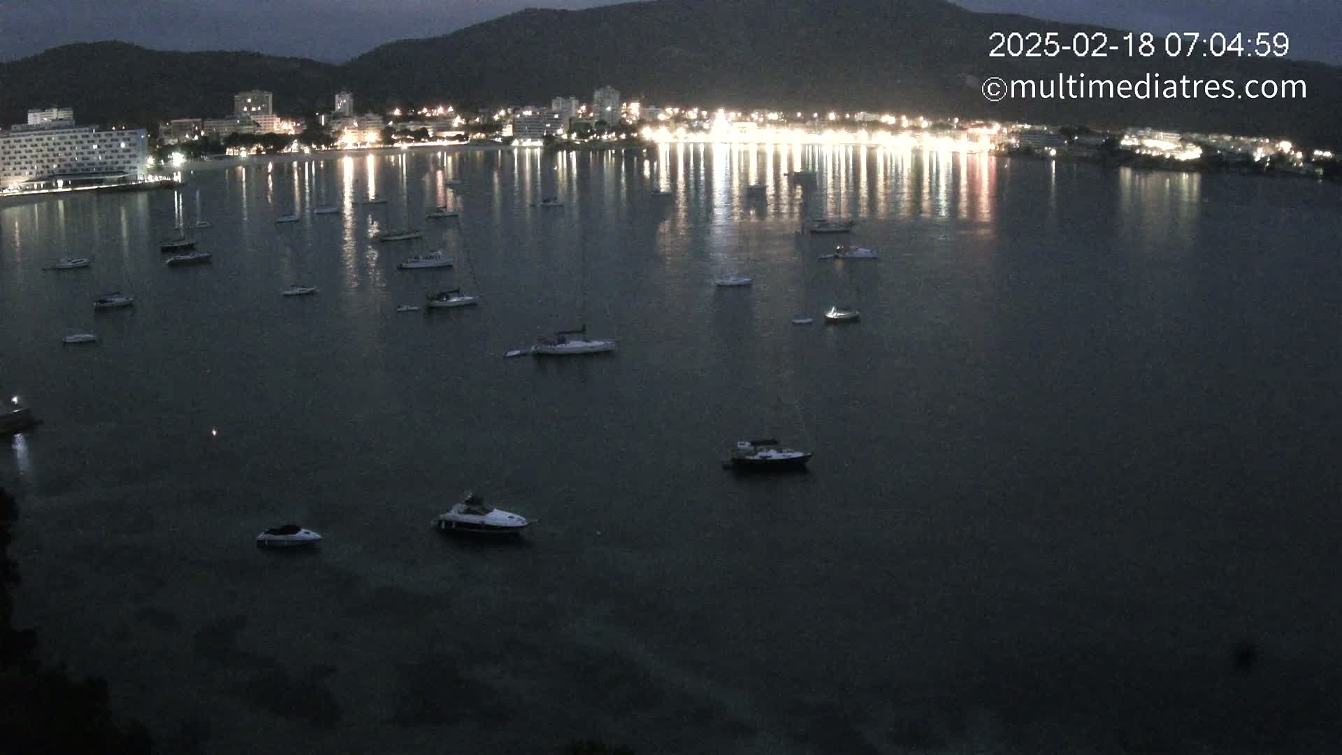 Numerous boats are moored in a dark, calm bay at night, illuminated by the lights of a town on the far shore.