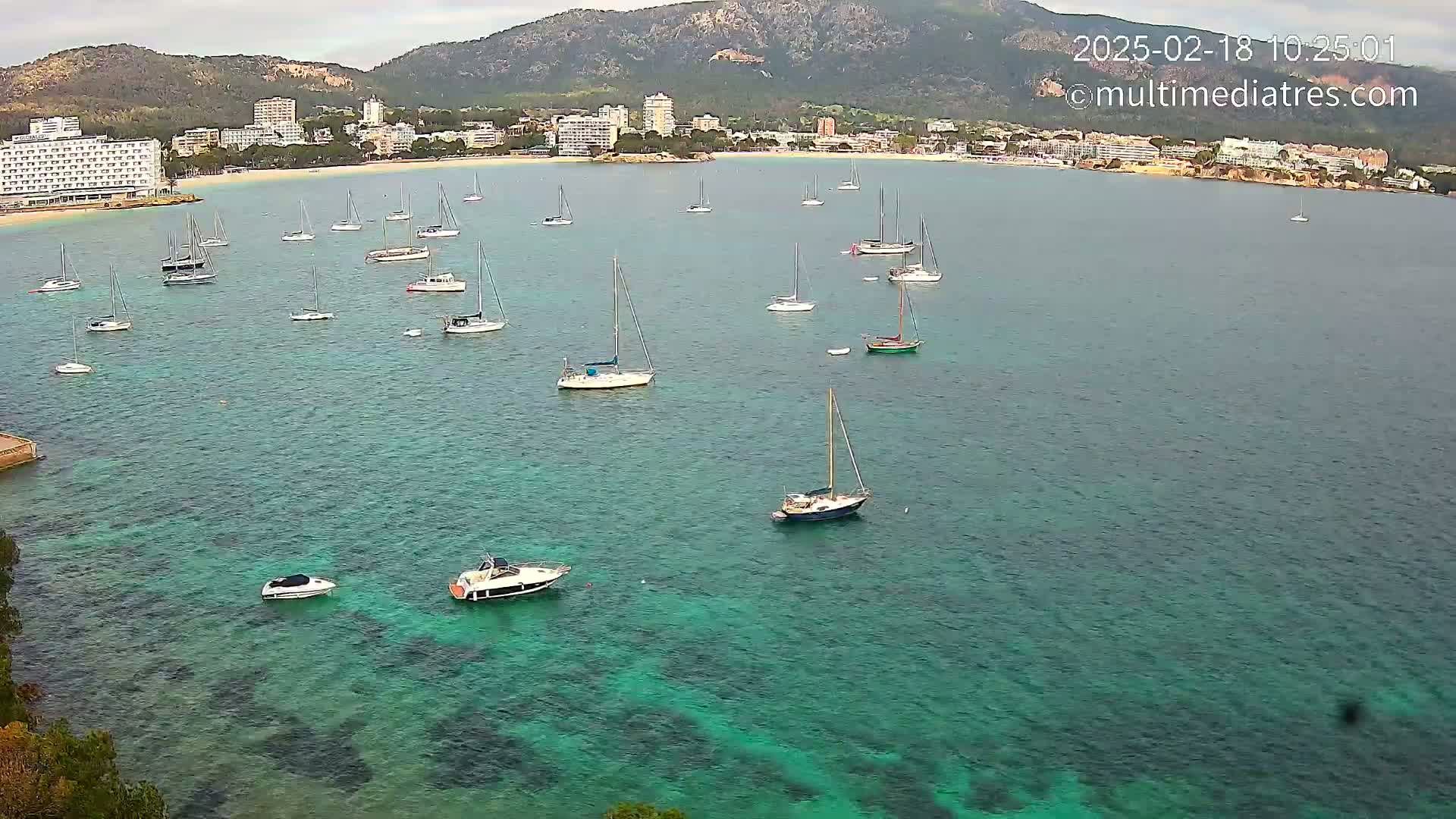 Numerous sailboats and motorboats are anchored in a calm, clear turquoise bay near a coastline with hotels and mountains under an overcast sky.