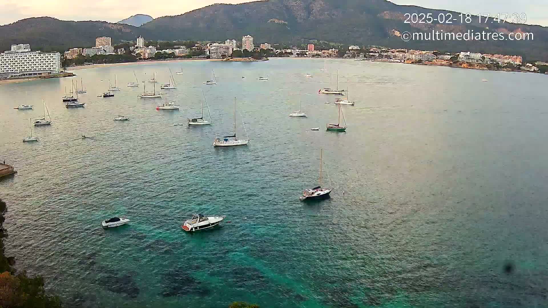 Numerous sailboats and motorboats are anchored in a calm, teal-colored bay near a coastline with buildings and mountains in the background under a partly cloudy sky.