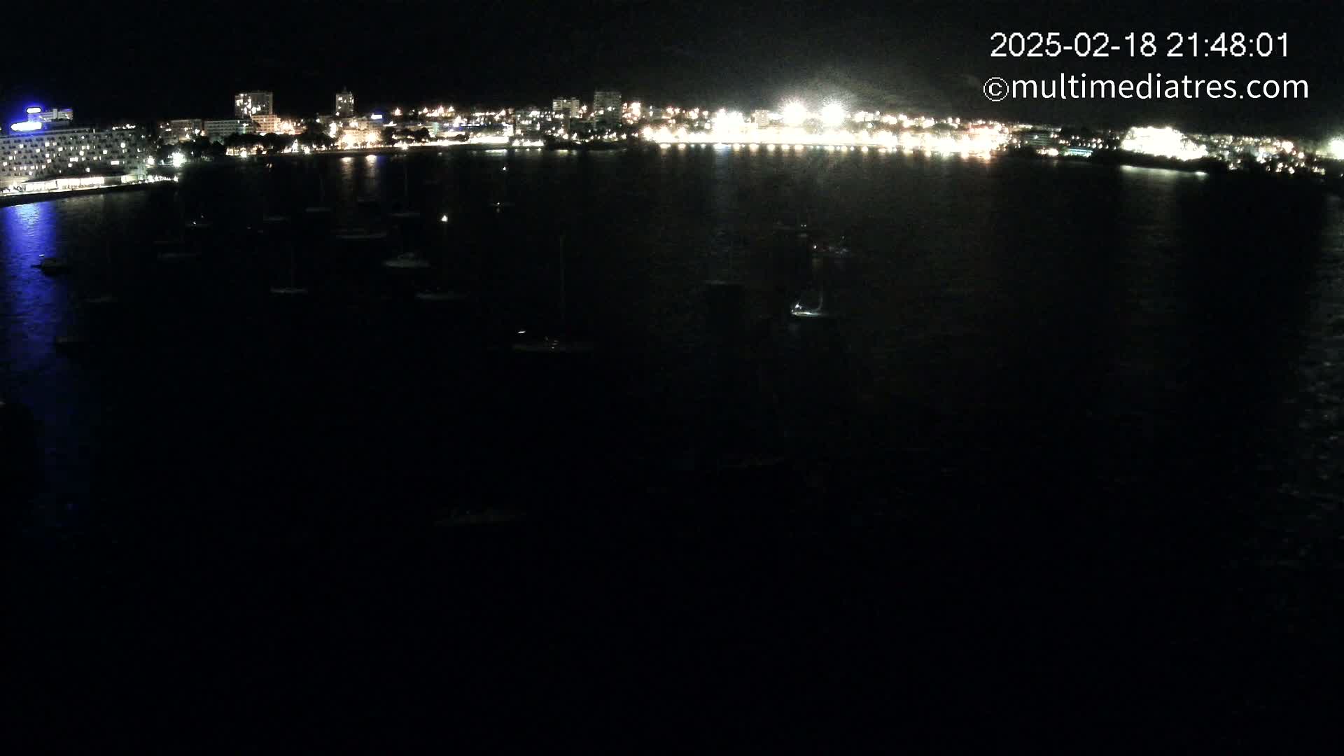 At night, a city's illuminated waterfront stretches across the horizon, with numerous small boats moored in the dark water.