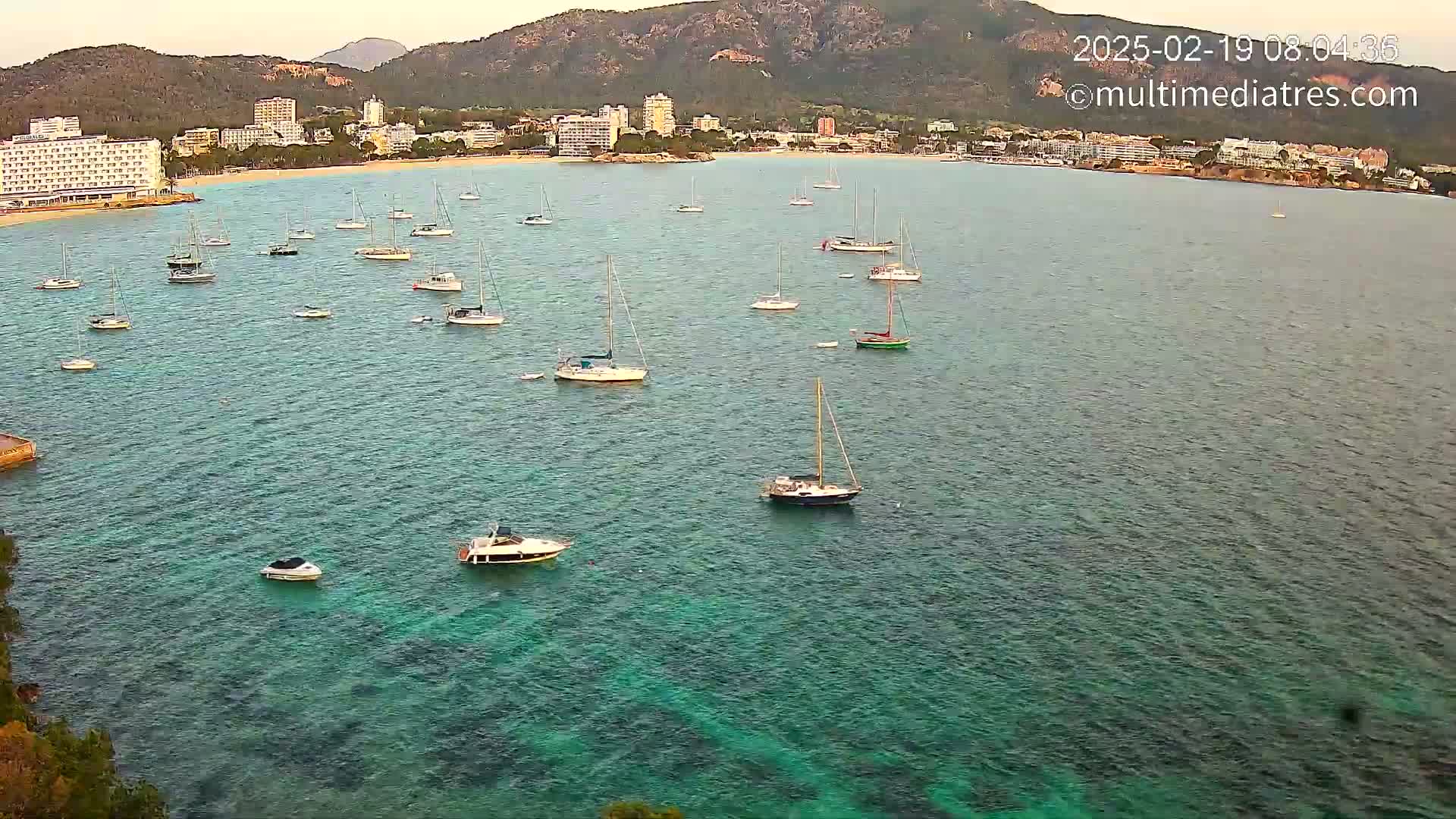 Numerous sailboats and motorboats are anchored in a calm, clear teal bay, with a town and mountains visible in the background.
