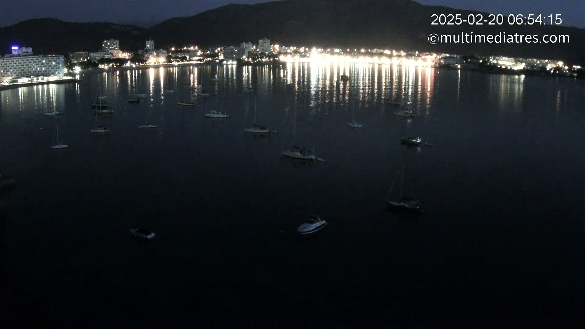 A dark, nighttime view of a calm bay with numerous boats moored, reflecting the lights of a city on a hill in the background.