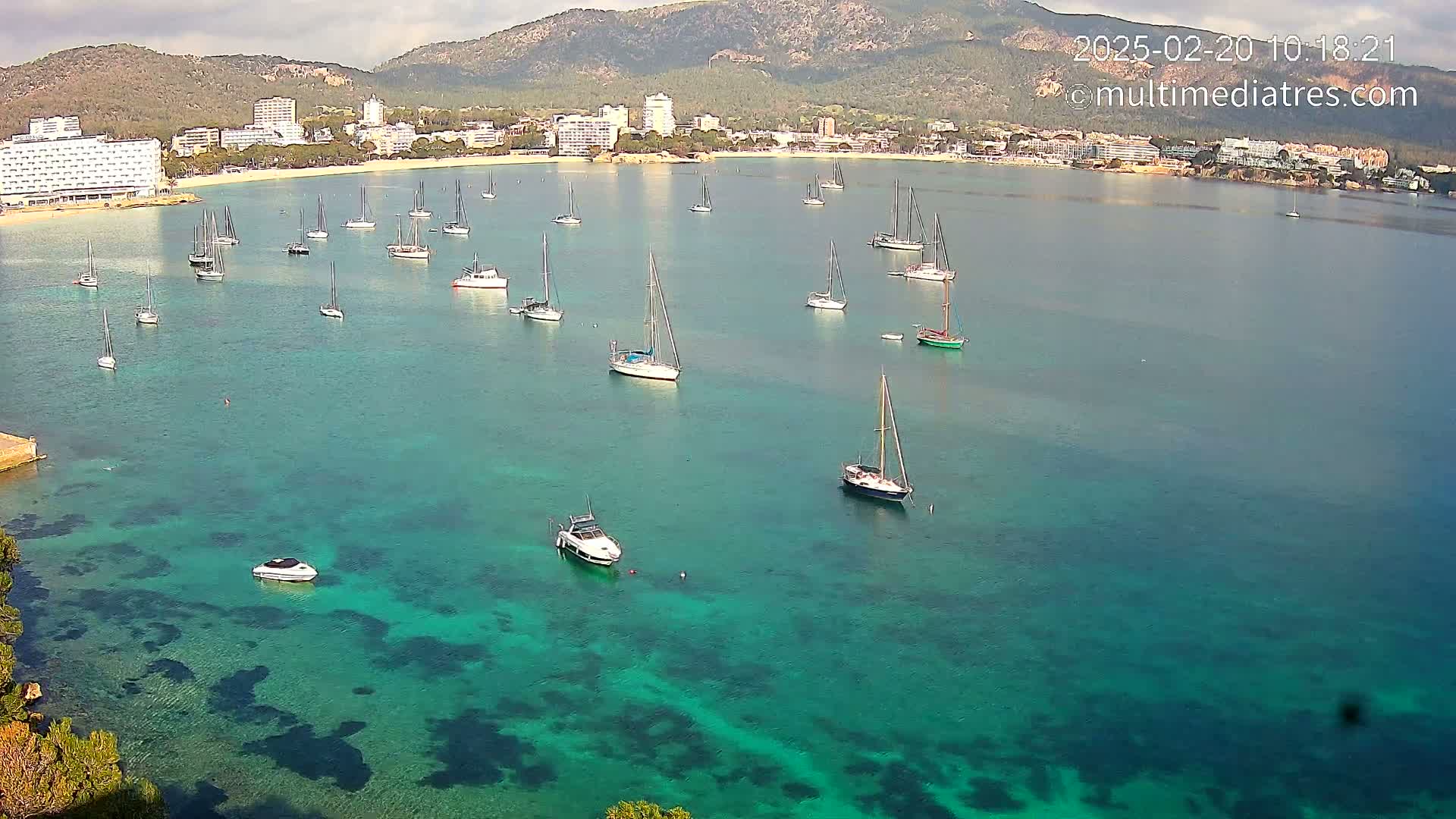 Numerous sailboats and motorboats are anchored in a calm, clear, teal bay near a coastline with hotels and mountains under a partly cloudy sky.