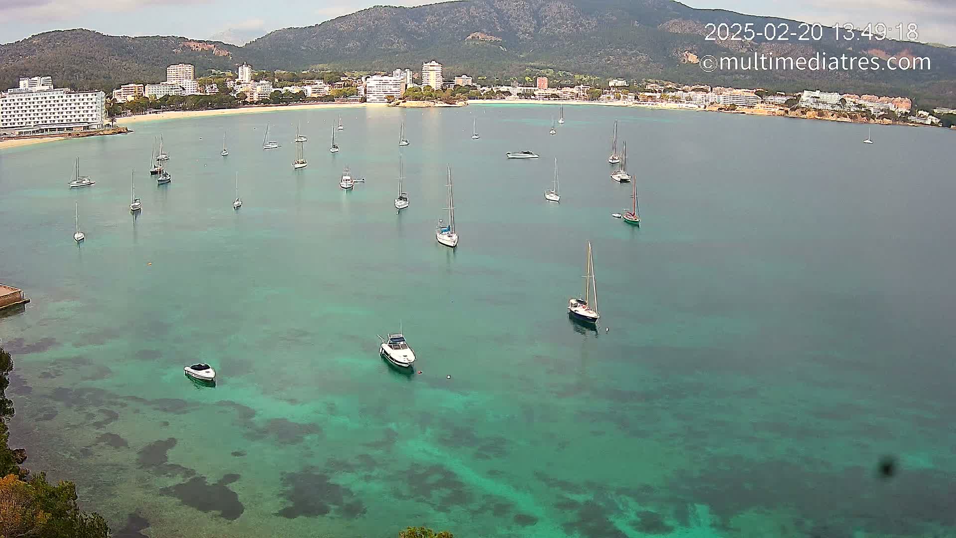 A calm, overcast day shows numerous sailboats and motorboats anchored in a teal-colored bay, with a sandy beach and hotels visible on the distant shore beneath a mountain range.