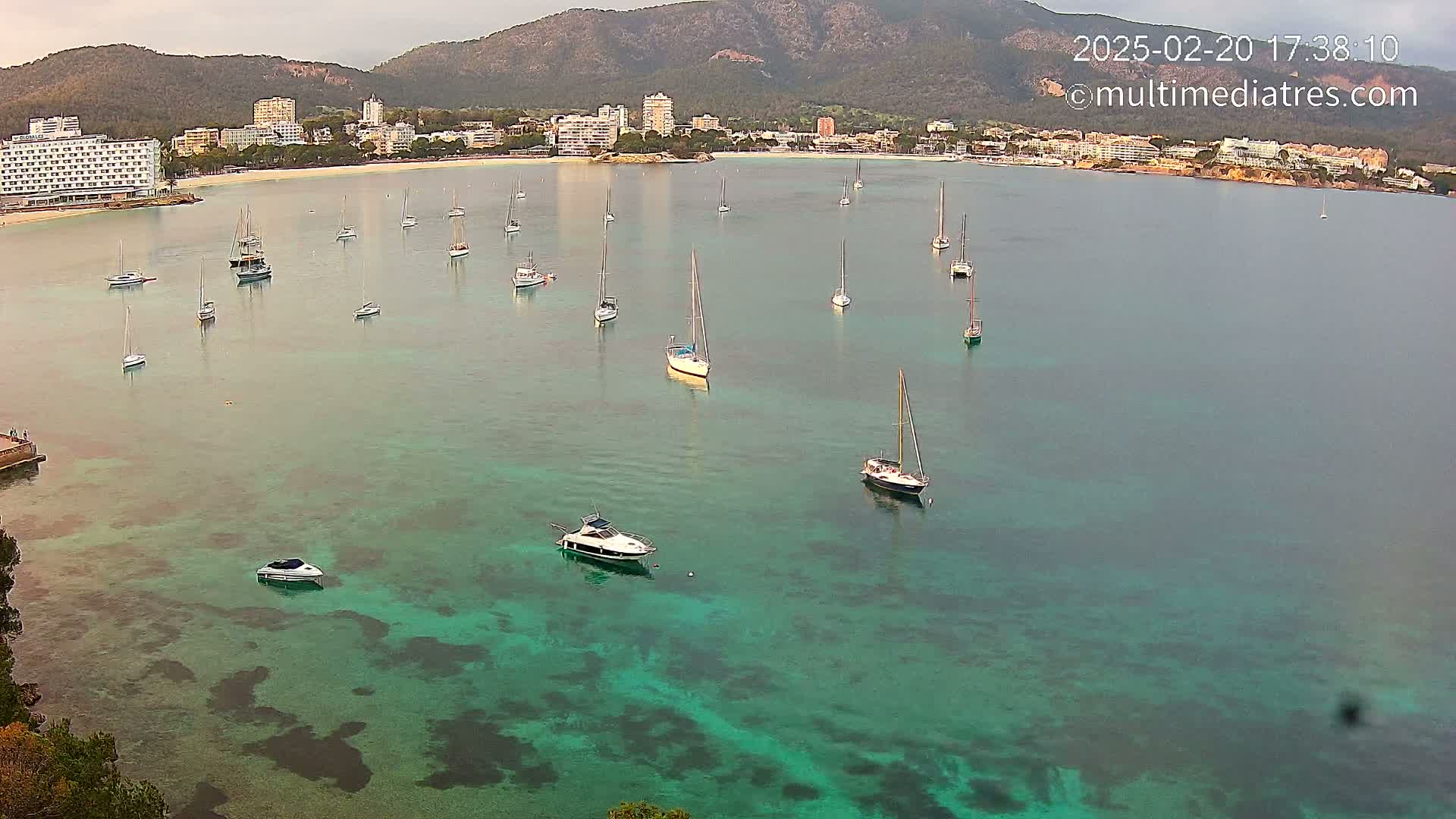 A calm, clear teal bay with numerous sailboats and motorboats anchored near a shoreline of hotels and buildings backed by mountains.