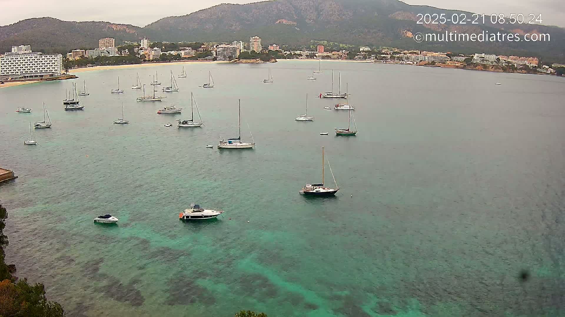 Numerous sailboats and motorboats are anchored in a calm, teal-colored bay, with a town and mountains visible in the background under an overcast sky.