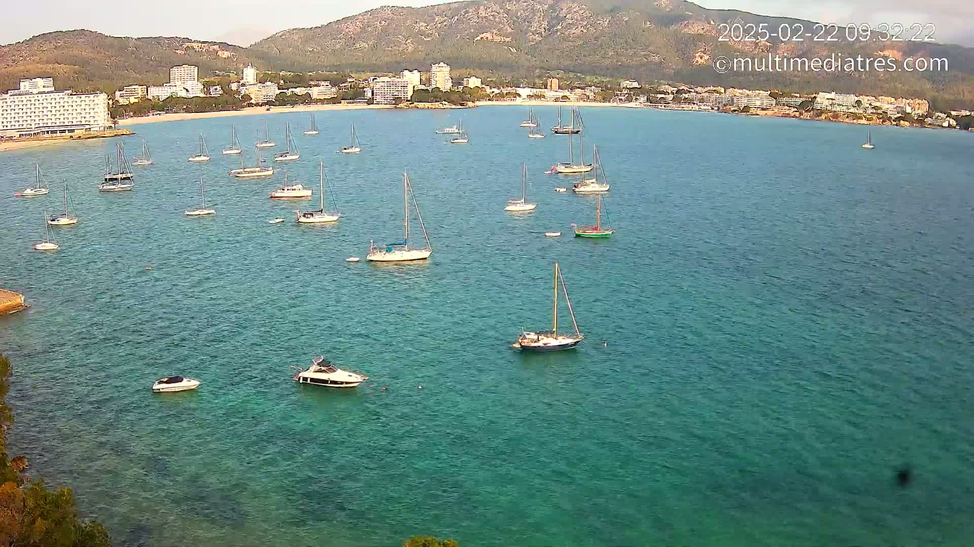 Numerous sailboats and motorboats are anchored in a calm, clear turquoise bay, with a shoreline of hotels and mountains in the background.