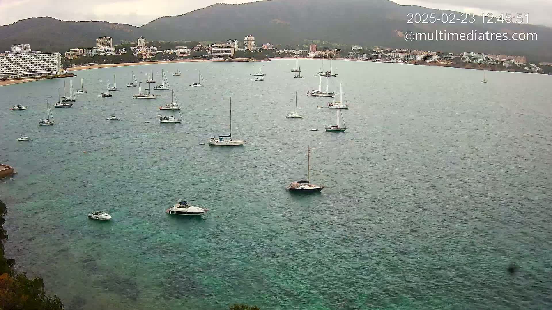 Numerous sailboats and motorboats are anchored in a calm, teal-colored bay, with a town and mountains visible in the background under an overcast sky.