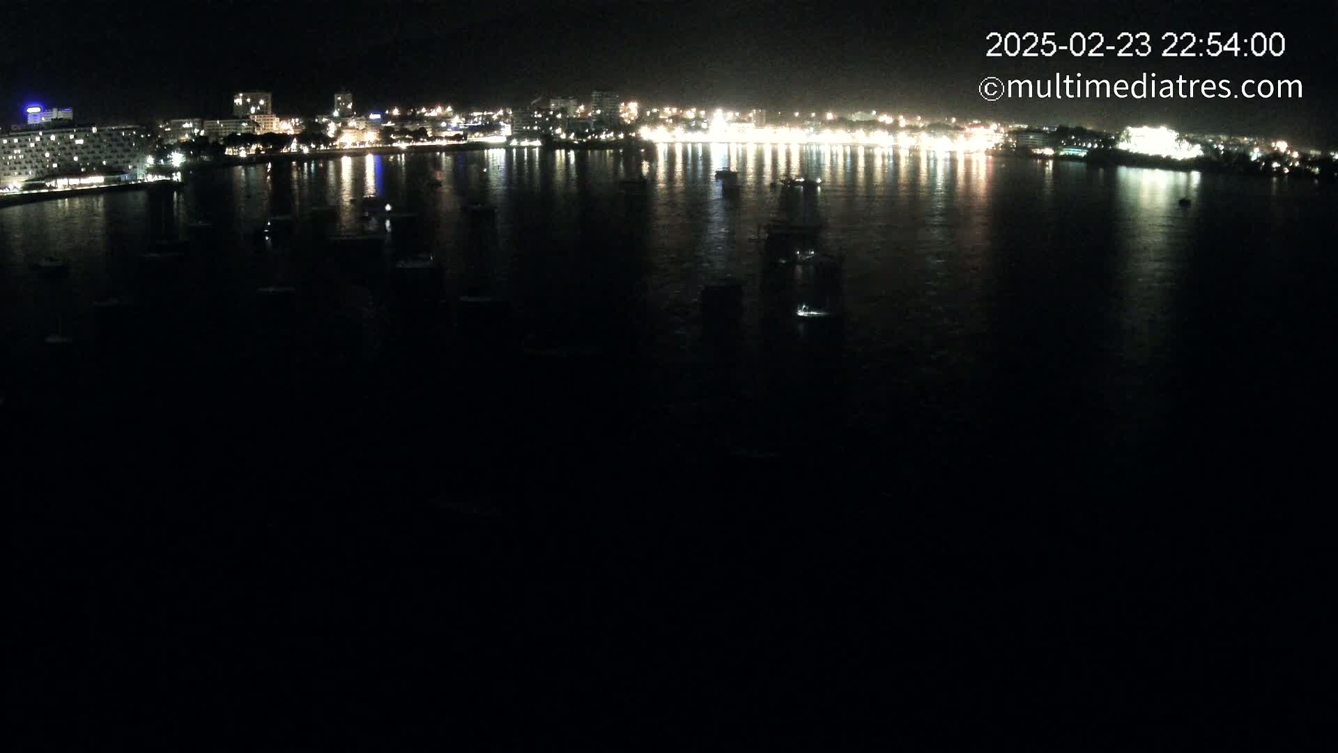 At night, a body of water reflects the lights of a city skyline and several boats are visible on the water.