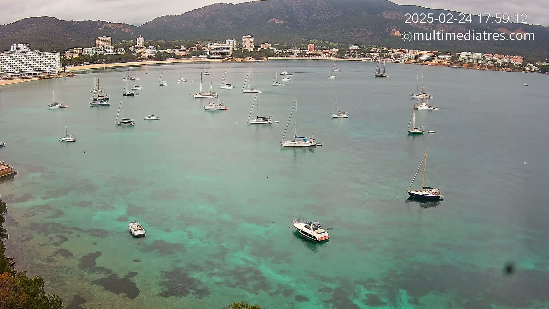 A calm, overcast day shows numerous sailboats and motorboats anchored in a turquoise bay, with a shoreline of hotels and mountains in the background.