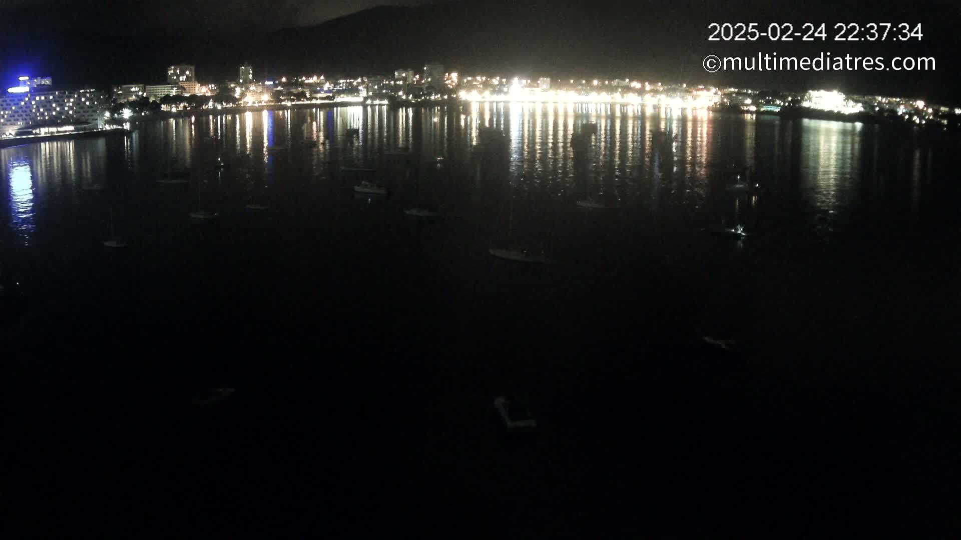 A nighttime view of a calm body of water with several boats and a brightly lit city reflected on its surface.