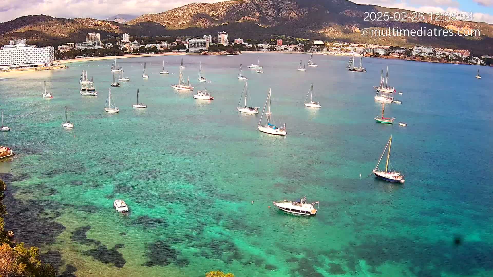 Numerous sailboats are anchored in a calm, clear, turquoise bay near a coastline with hotels and mountains under a partly cloudy sky.