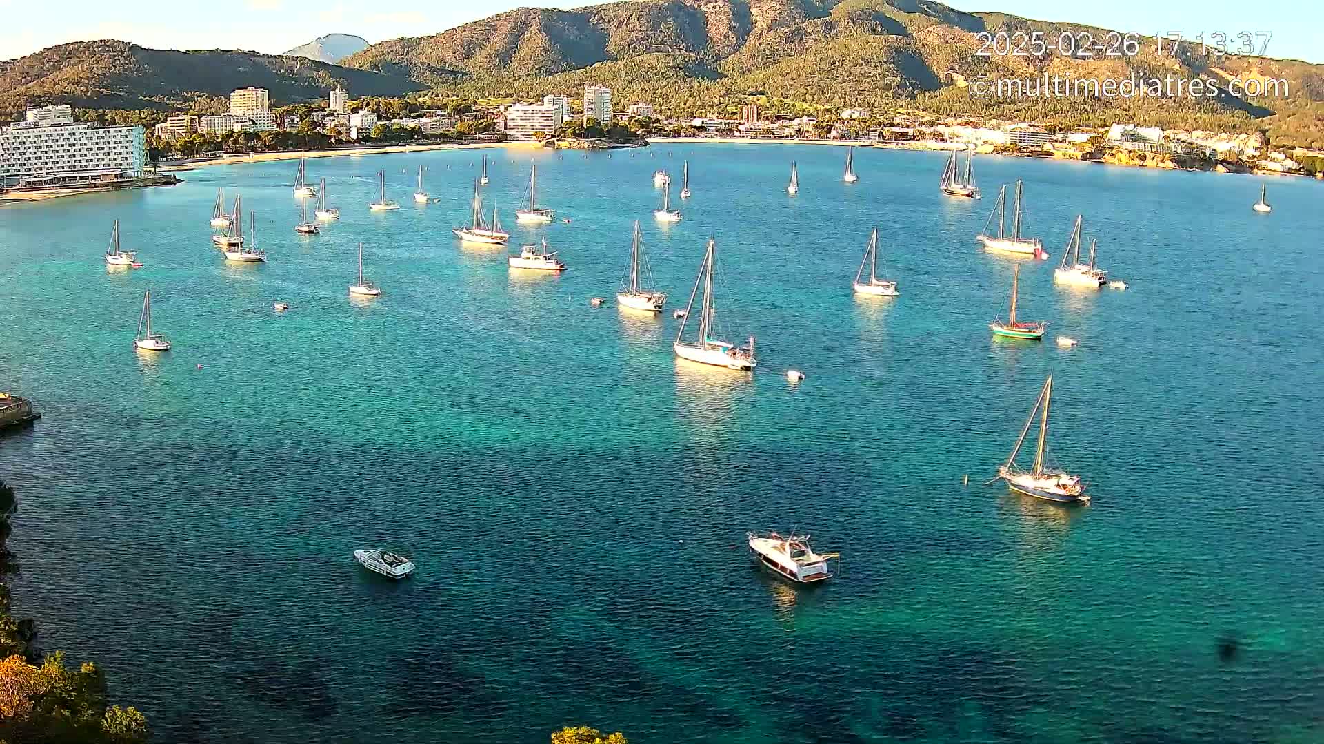 Numerous sailboats are anchored in a calm, clear turquoise bay, with a mountainous coastline and buildings in the background under a mostly sunny sky.