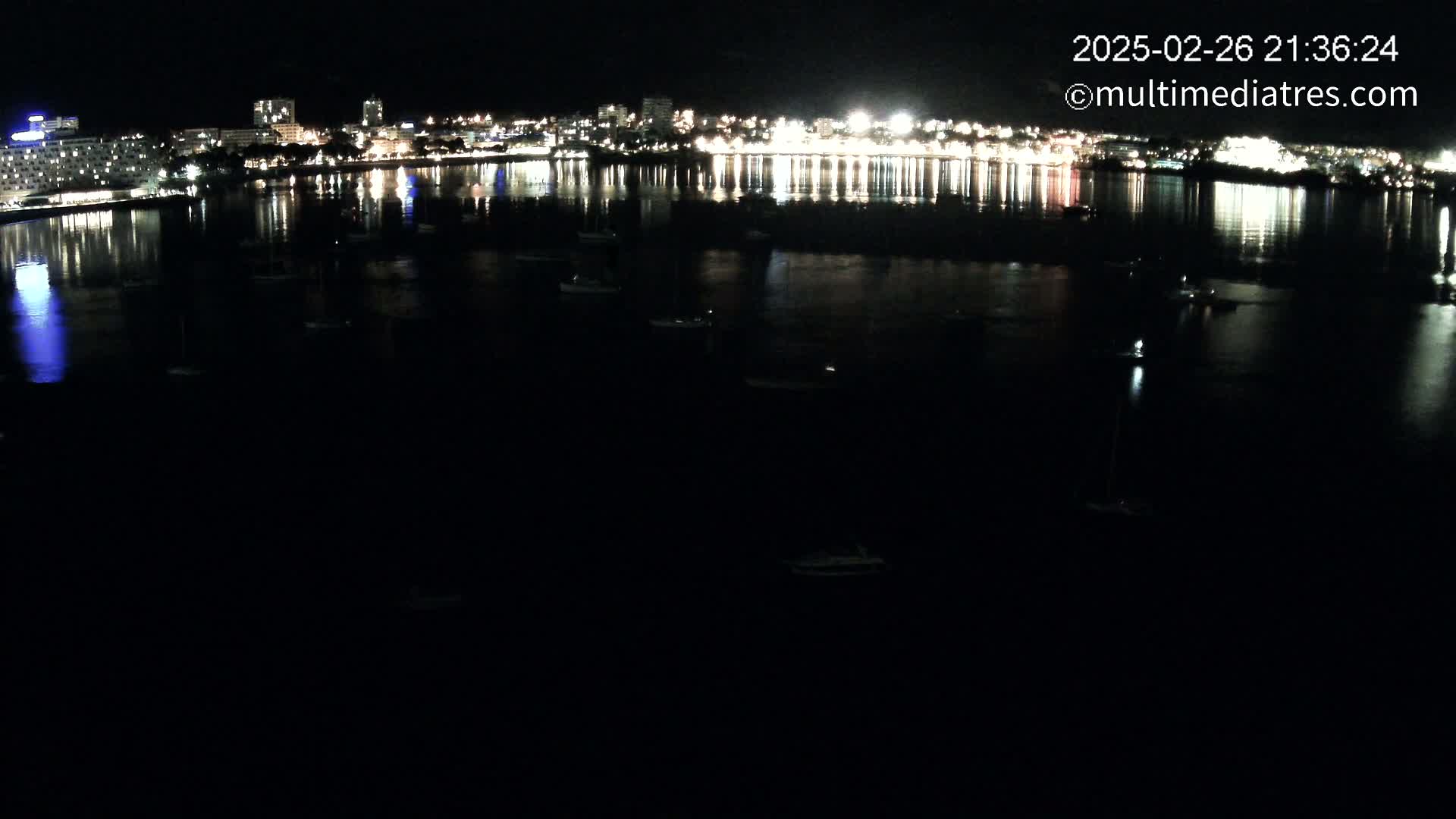 At night, a calm body of water reflects the lights of a city skyline dotted with several small boats.