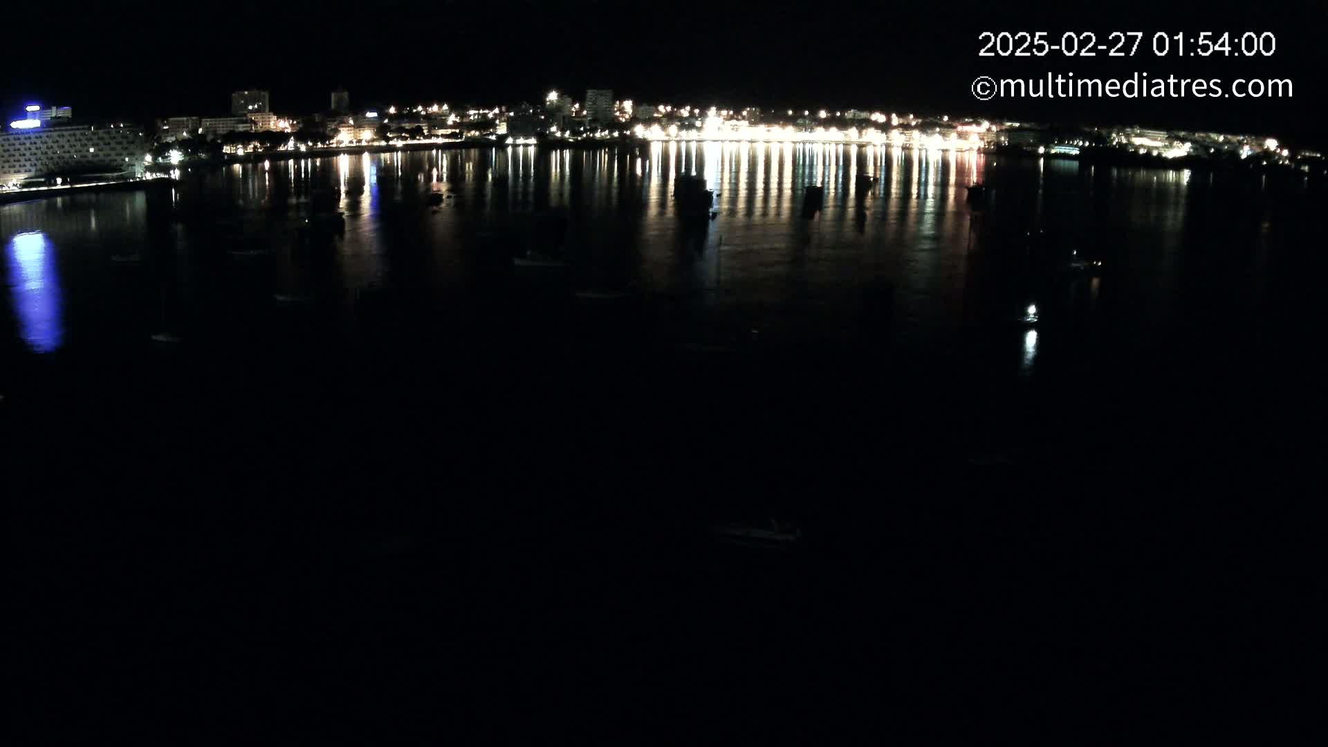 A nighttime view shows a calm body of water reflecting the lights of a city skyline, with a few small boats scattered across the dark water.