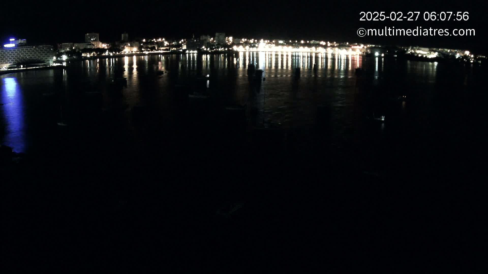 A nighttime view of a city's illuminated waterfront, reflected in a calm body of water, with several boats visible.