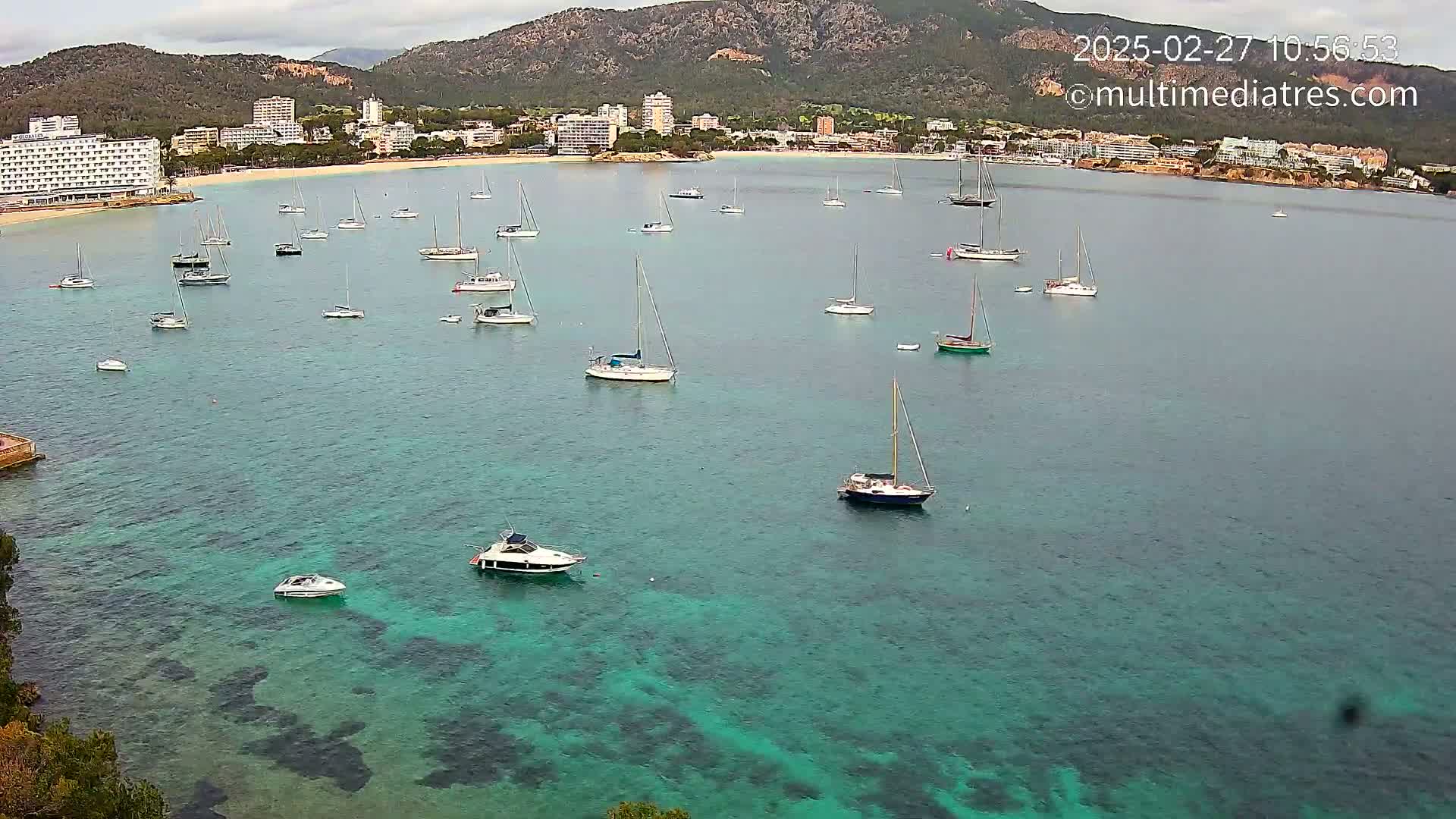 Numerous sailboats and motorboats are anchored in a calm, teal-colored bay near a beach and hotels situated against a mountainous backdrop under an overcast sky.