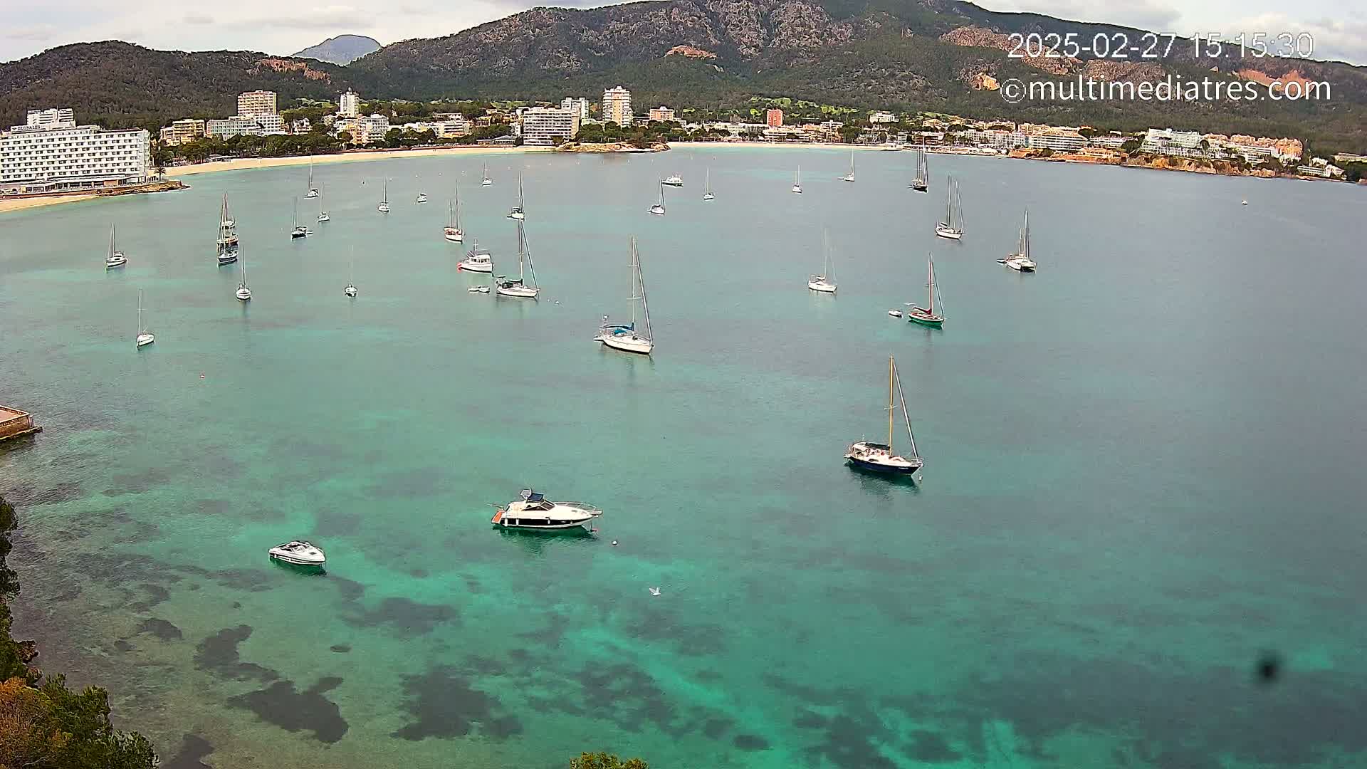 Numerous sailboats and motorboats are anchored in a calm, teal bay near a shoreline with hotels and mountains under a cloudy sky.