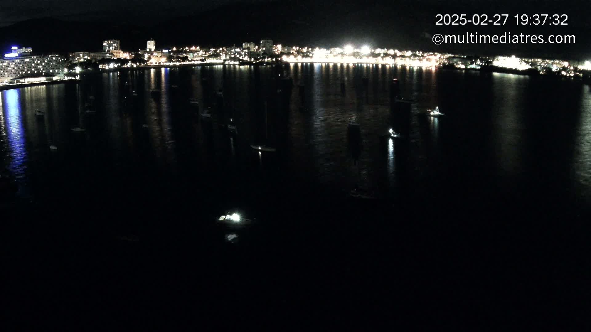 Numerous boats are moored in a dark body of water at night in front of a brightly lit city skyline.