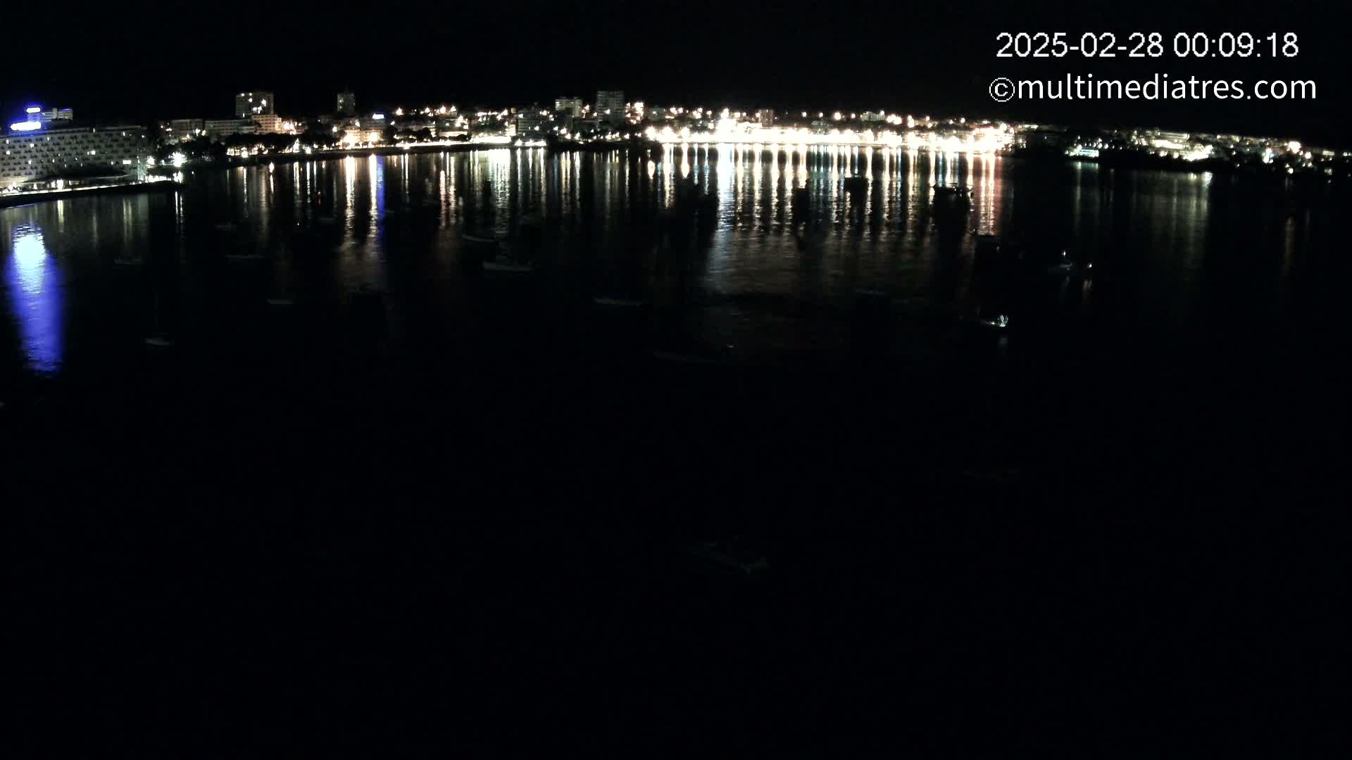 A nighttime view of a calm body of water reflecting the lights of a city skyline across its surface, with several small boats visible.