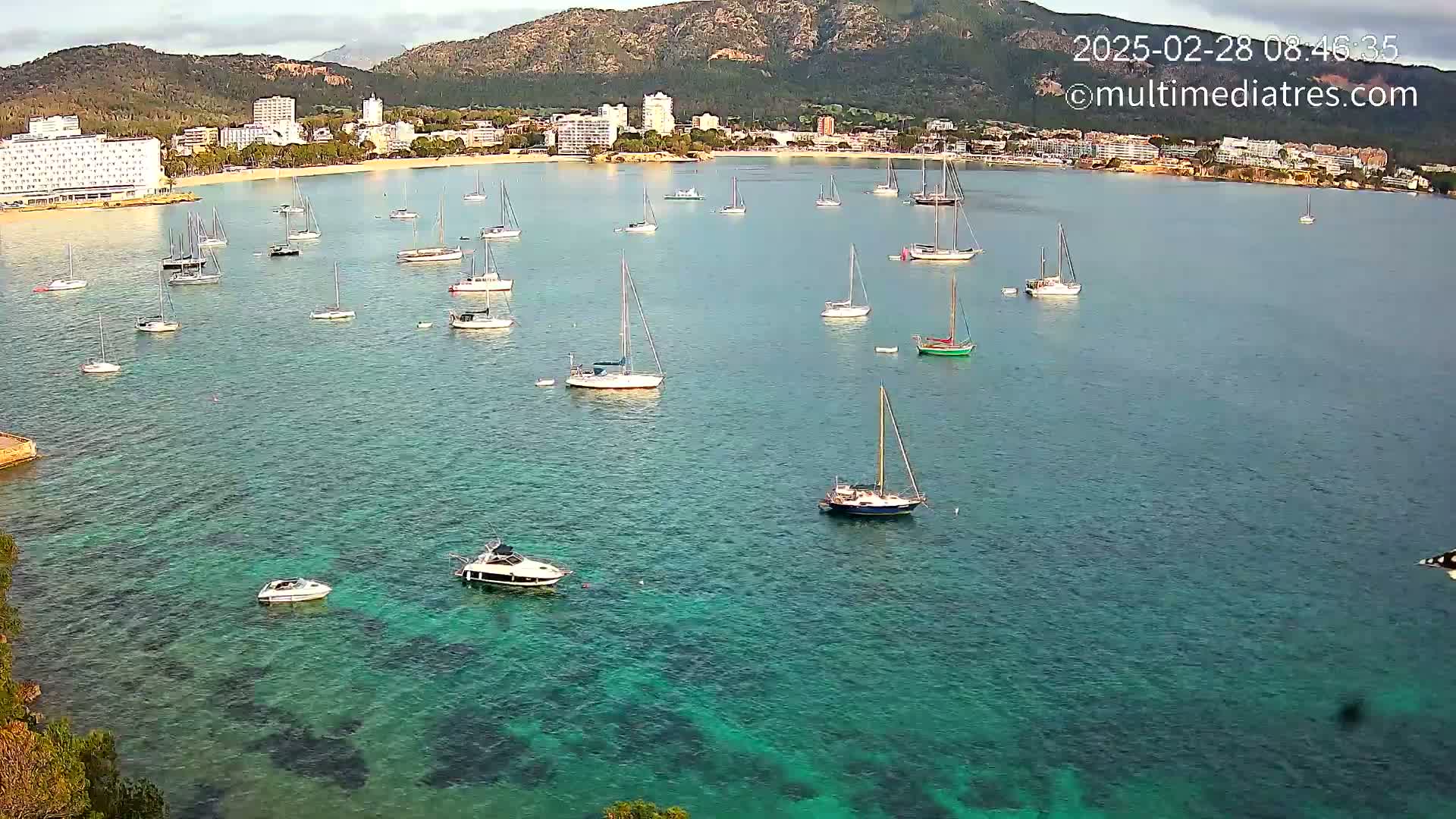 Numerous sailboats and motorboats are anchored in a calm, clear turquoise bay near a coastline with hotels and mountains in the background under a mostly clear sky.