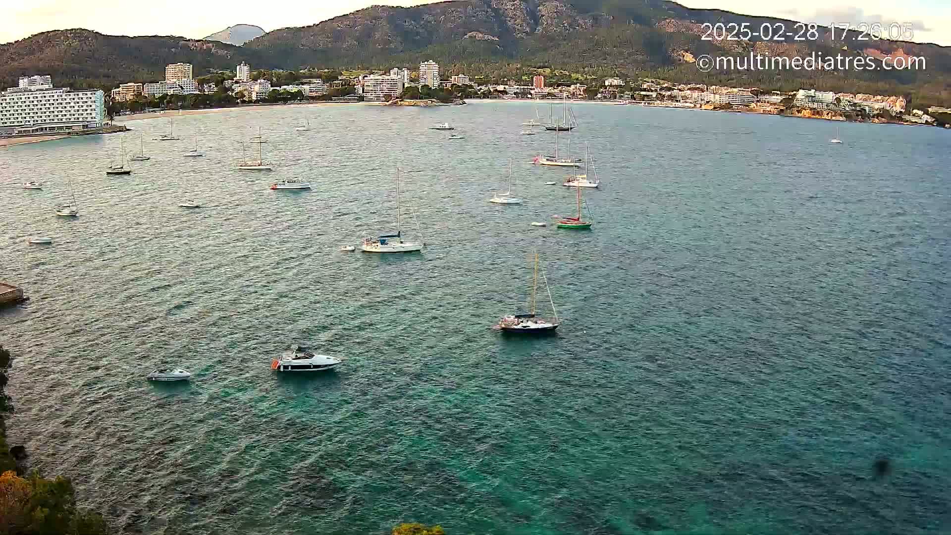 Numerous sailboats and motorboats are anchored in a calm, clear teal sea near a coastline with hotels and mountains in the background.