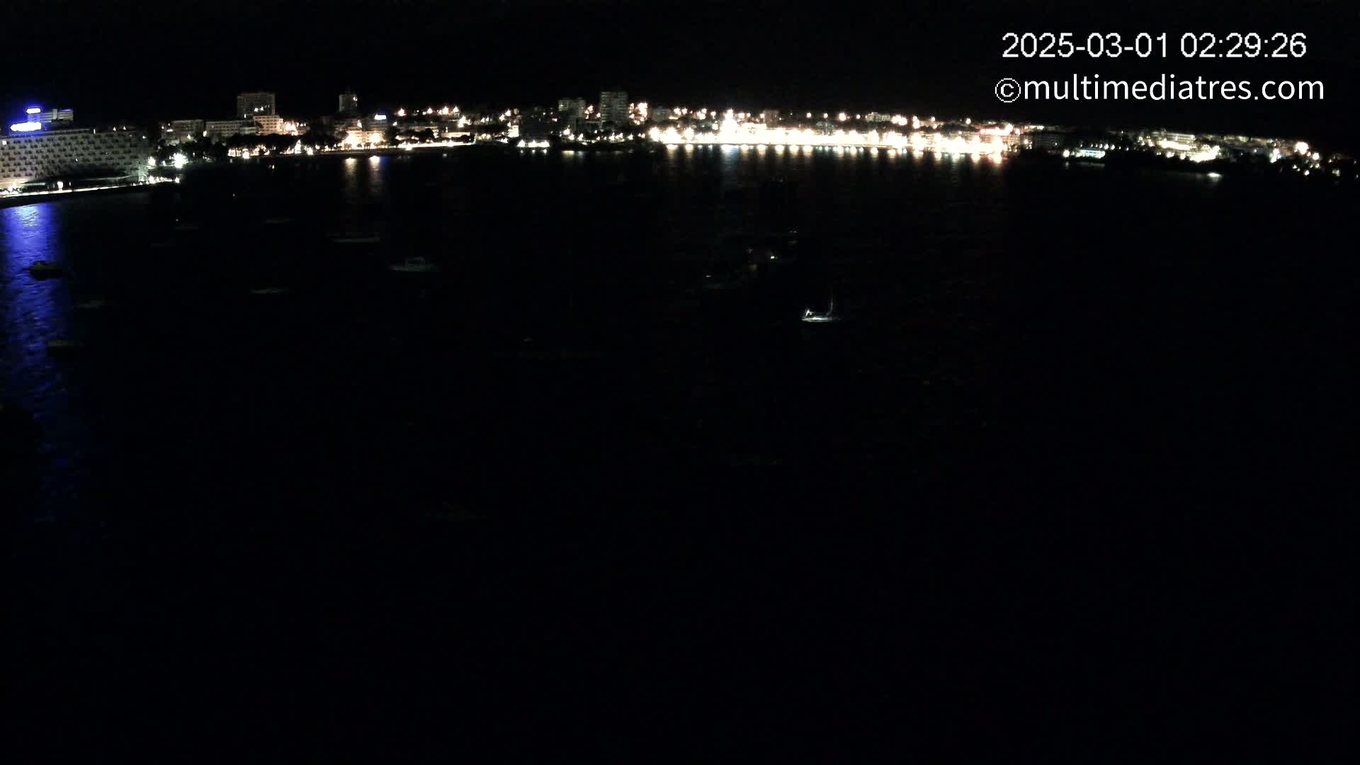 A nighttime view of a city's illuminated waterfront, with several small boats scattered on dark water.