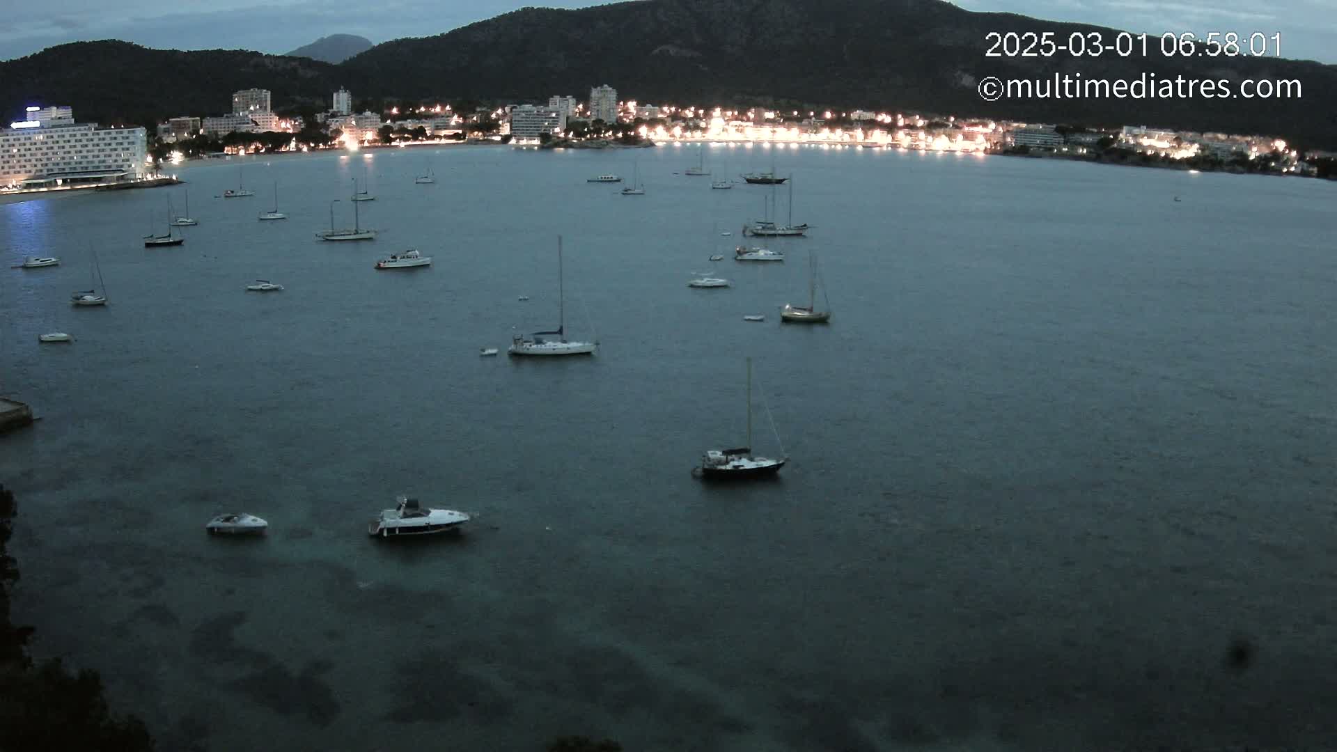 Many sailboats and motorboats are moored in a calm bay at twilight, with a town's lights visible on the shore against a dark mountain backdrop.