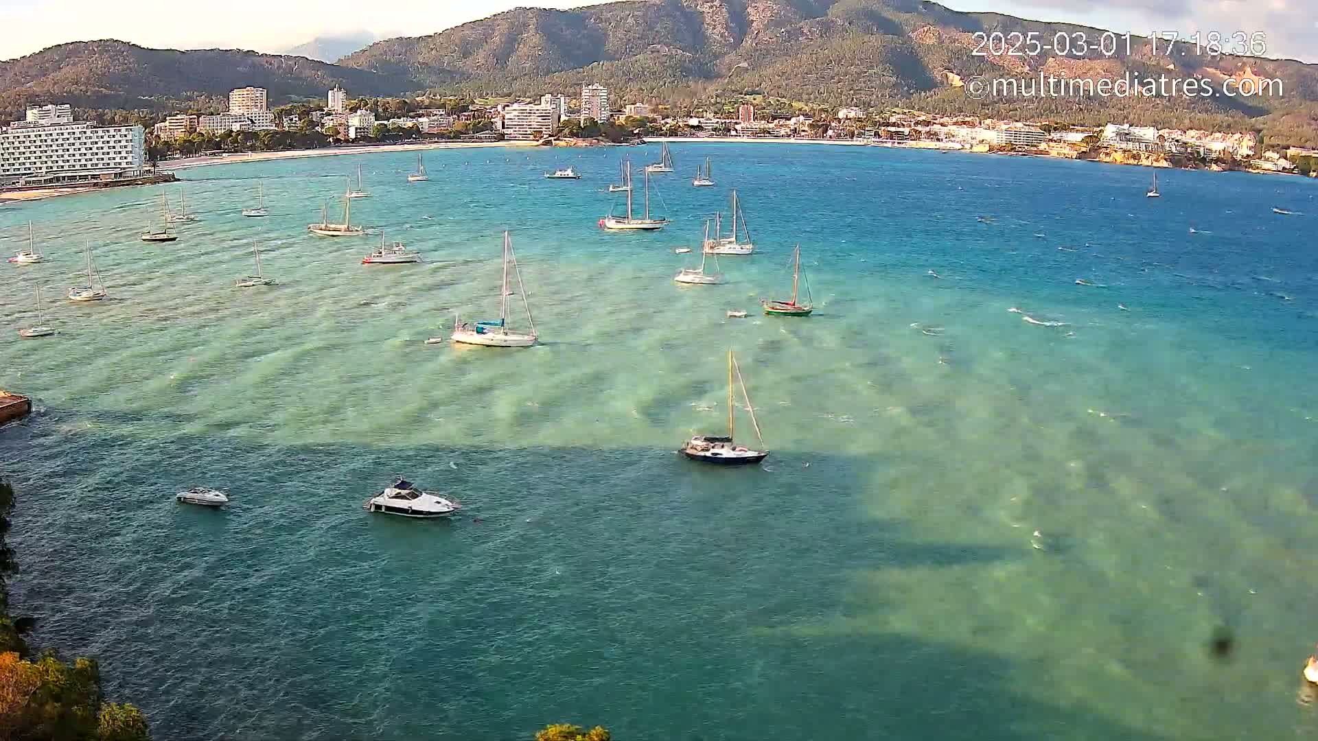 Numerous sailboats and motorboats are anchored in a calm, clear bay next to a coastline with hotels and mountains in the background.
