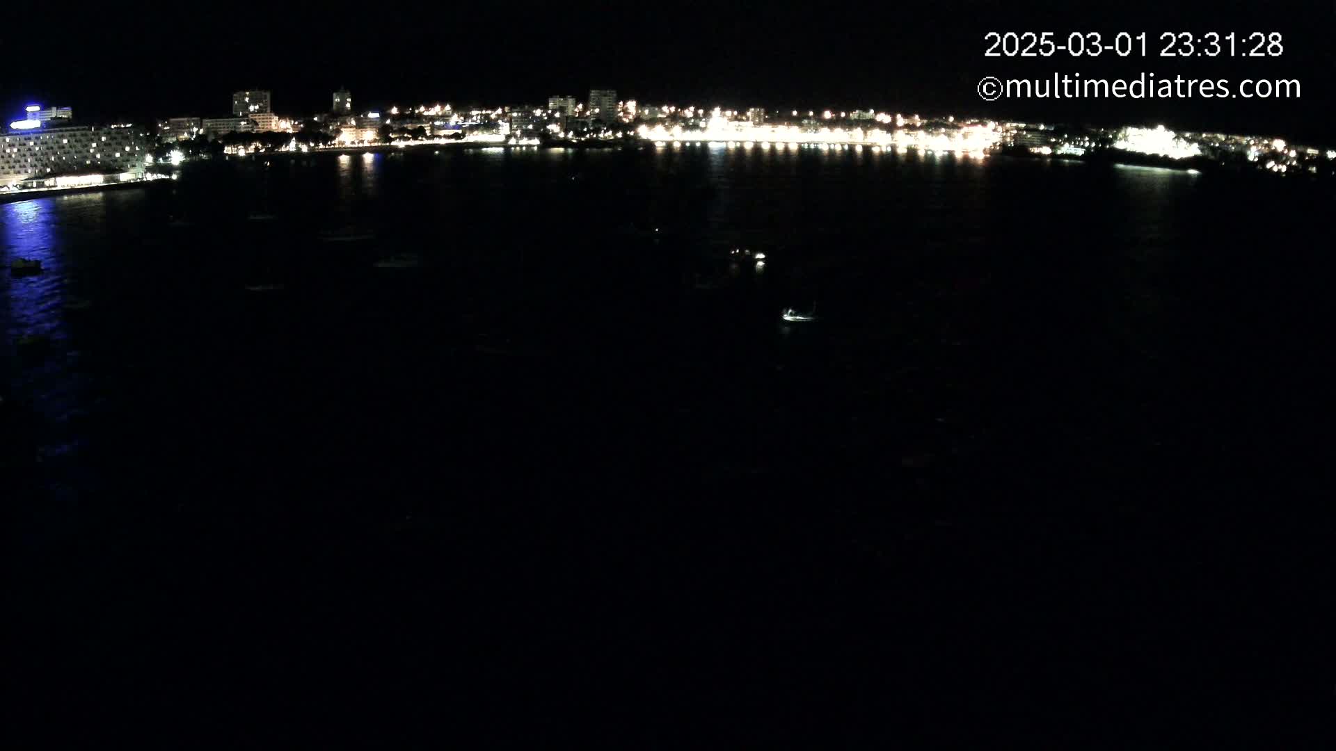 A nighttime view of a city's illuminated waterfront, featuring several buildings and a dark body of water with a few small lights scattered across it.
