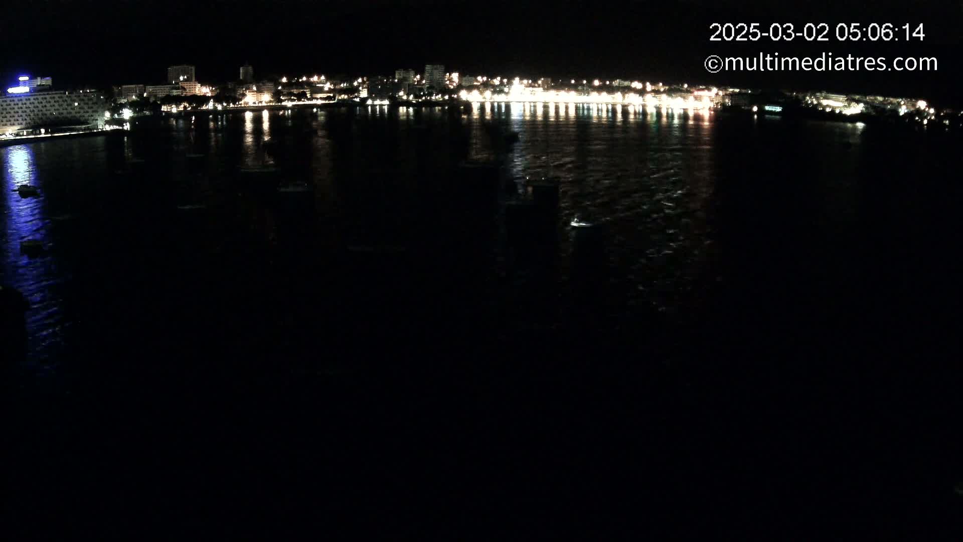 At night, a dark body of water reflects the lights of a city skyline and several small boats.