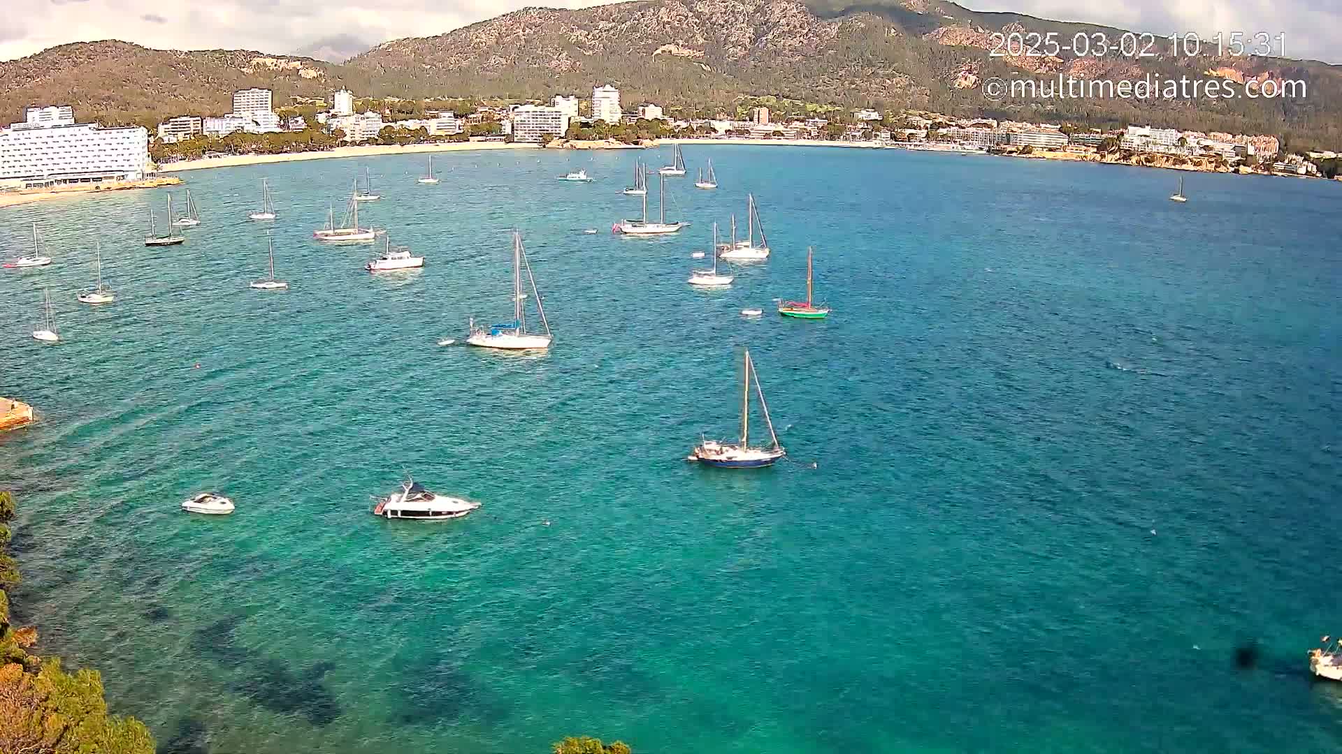 A calm, clear turquoise body of water dotted with sailboats and motorboats, sits in front of a coastline with buildings and a mountain range under a partly cloudy sky.