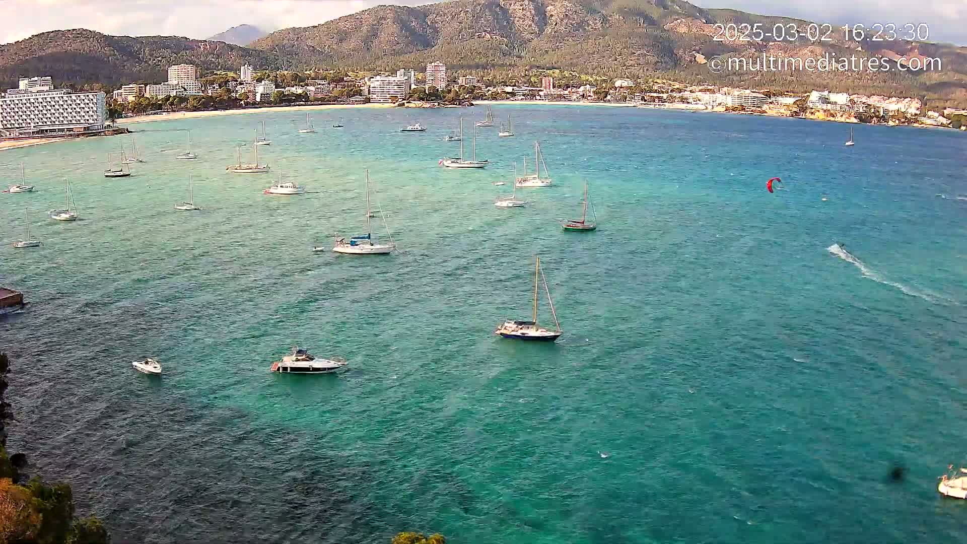 Numerous sailboats and motorboats are anchored in a calm, teal-colored bay, with a kitesurfer visible in the distance, against a backdrop of mountains and a coastal town.