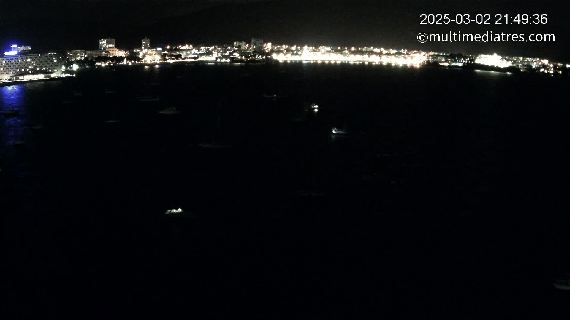A nighttime, aerial view shows a city's illuminated shoreline at night, with numerous small boats scattered across a dark body of water.