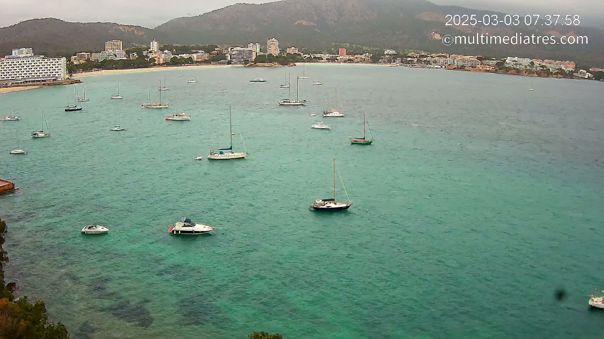 Numerous sailboats and motorboats are anchored in a calm, teal-colored bay under an overcast sky, with a shoreline of buildings and hills in the background.