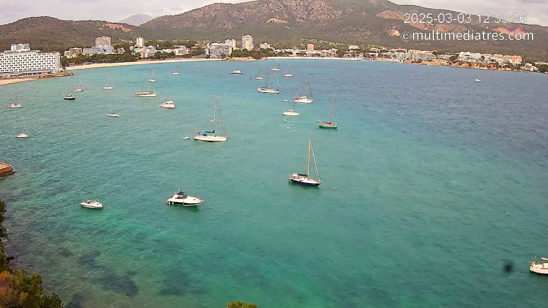Numerous sailboats and motorboats are anchored in a calm, teal-colored bay near a coastline with hotels and mountains under an overcast sky.