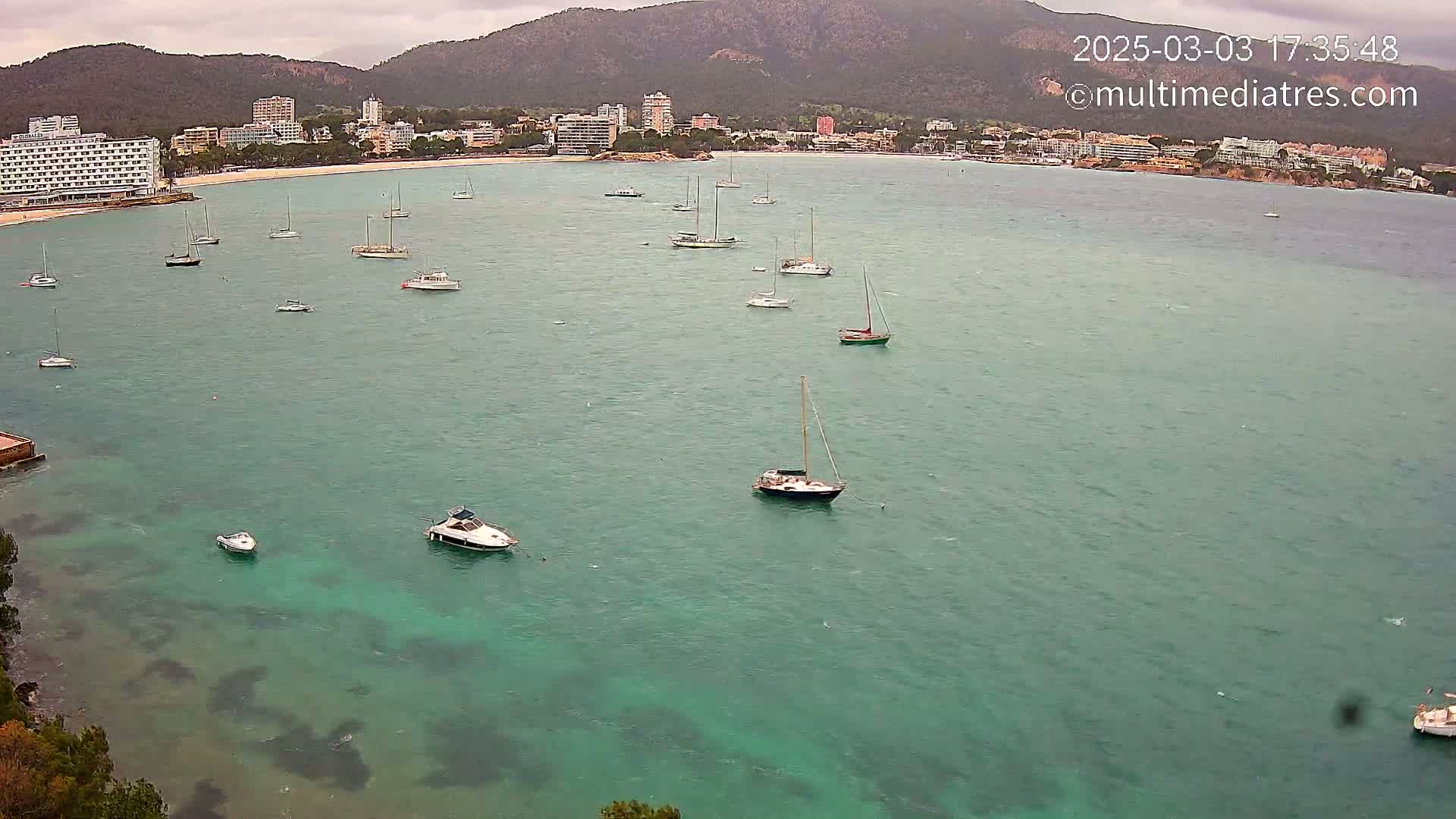 Numerous sailboats and motorboats are anchored in a calm, teal-colored bay under an overcast sky, with a beach and buildings visible in the background.
