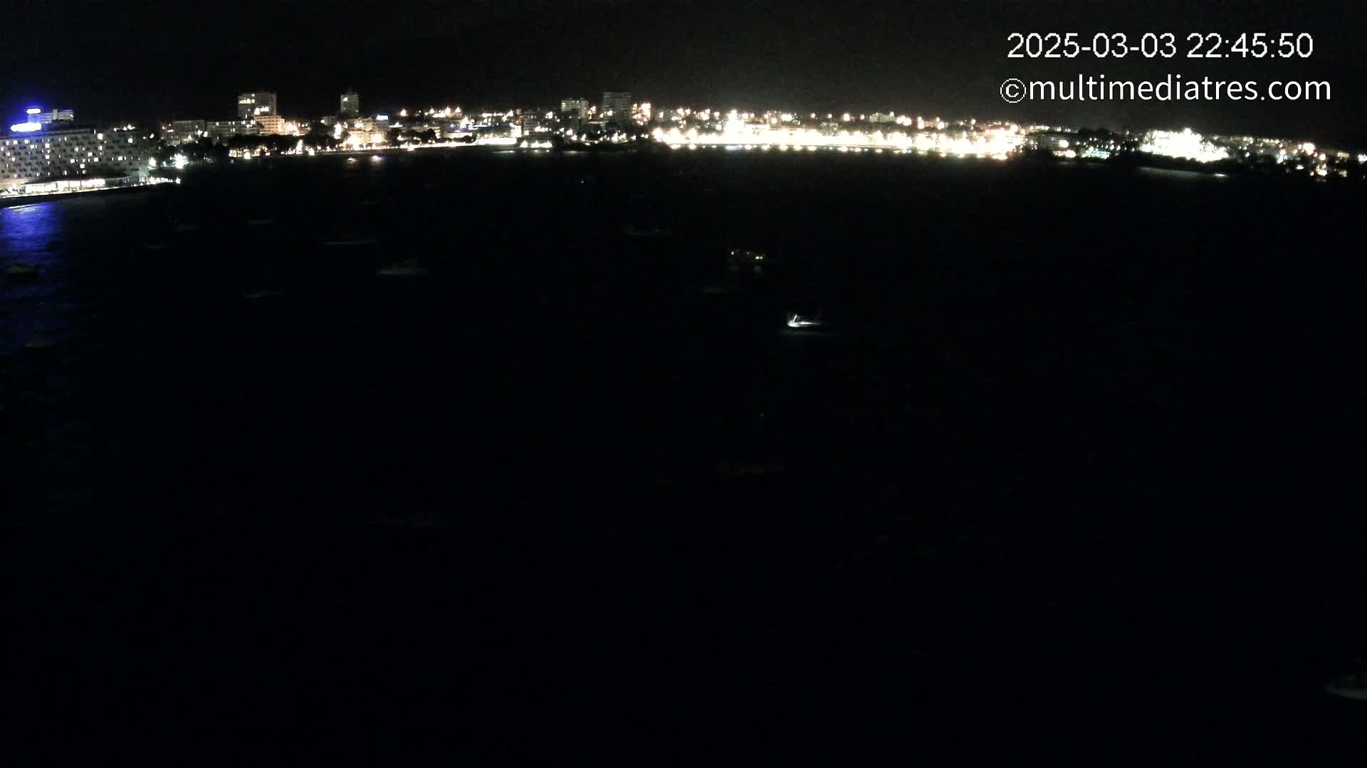 A nighttime, long-range view shows a dark body of water with a few small lights, in front of a city skyline brightly lit against the night sky.