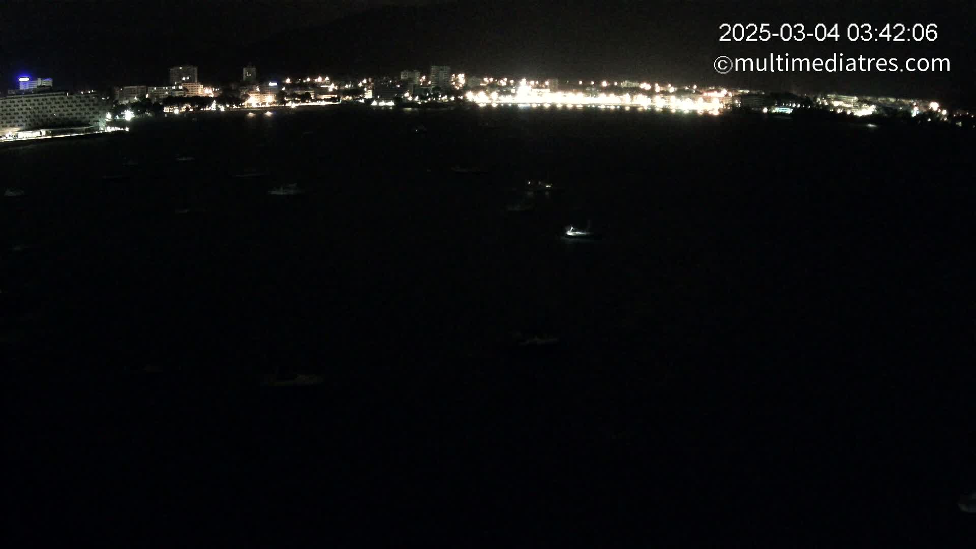 A dark, nighttime view of a city's illuminated waterfront with numerous small boats scattered across the dark water.