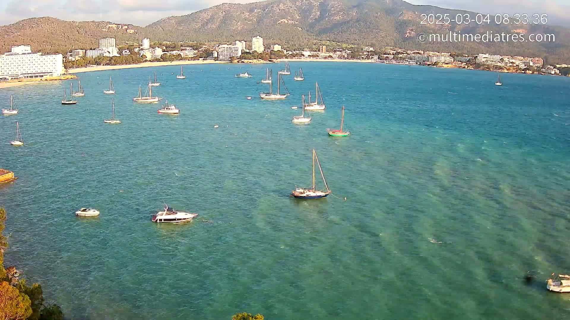 Numerous sailboats and motorboats are anchored in a calm, clear turquoise bay near a coastline with hotels and mountains in the background.